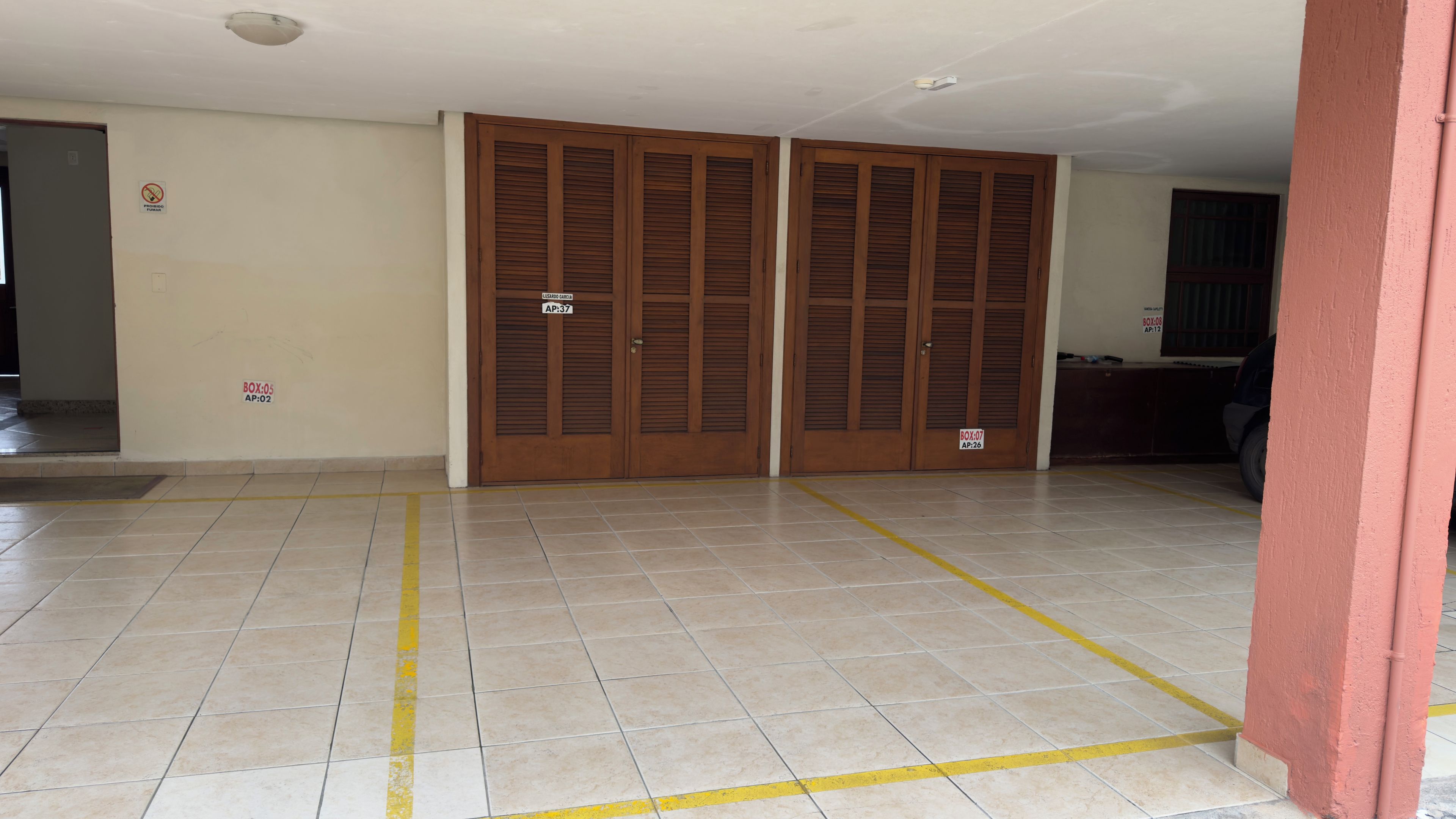 Um corredor com chão ladrilhado, portas de madeira com etiquetas e luminária fixada na parede.