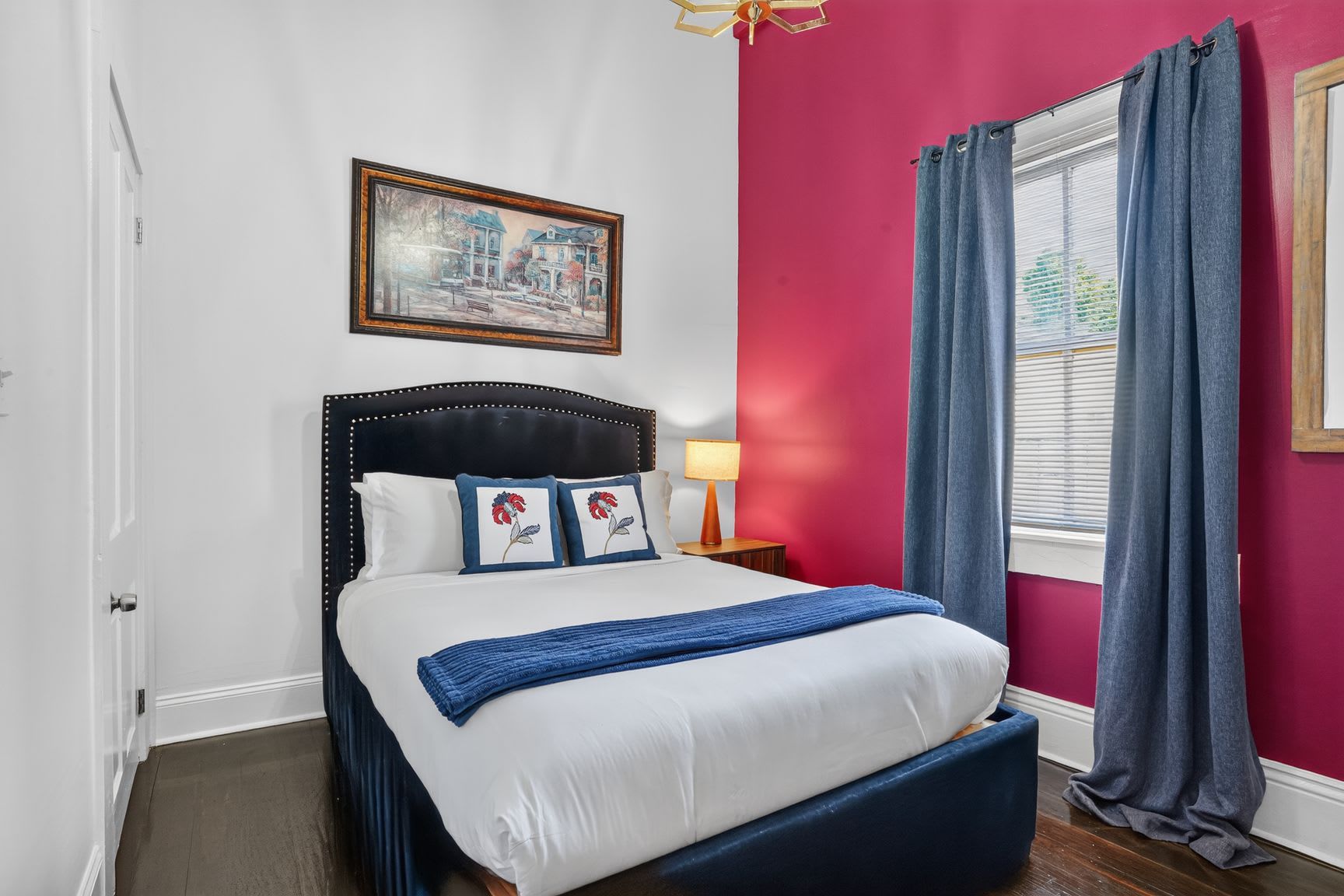 Une chambre à coucher avec un lit, une table de chevet, une lampe, une fenêtre avec des rideaux et un tableau au mur.