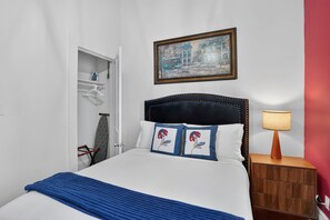 Un lit bien fait, recouvert d’une couverture bleue, avec une tête de lit ornée d’un coussin à motifs floraux, une lampe de chevet et un tableau accroché au-dessus du lit.