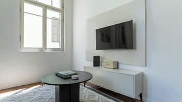 A modern living room with a round table, a flat-screen TV mounted on the wall, a white cabinet, and a window with blinds.