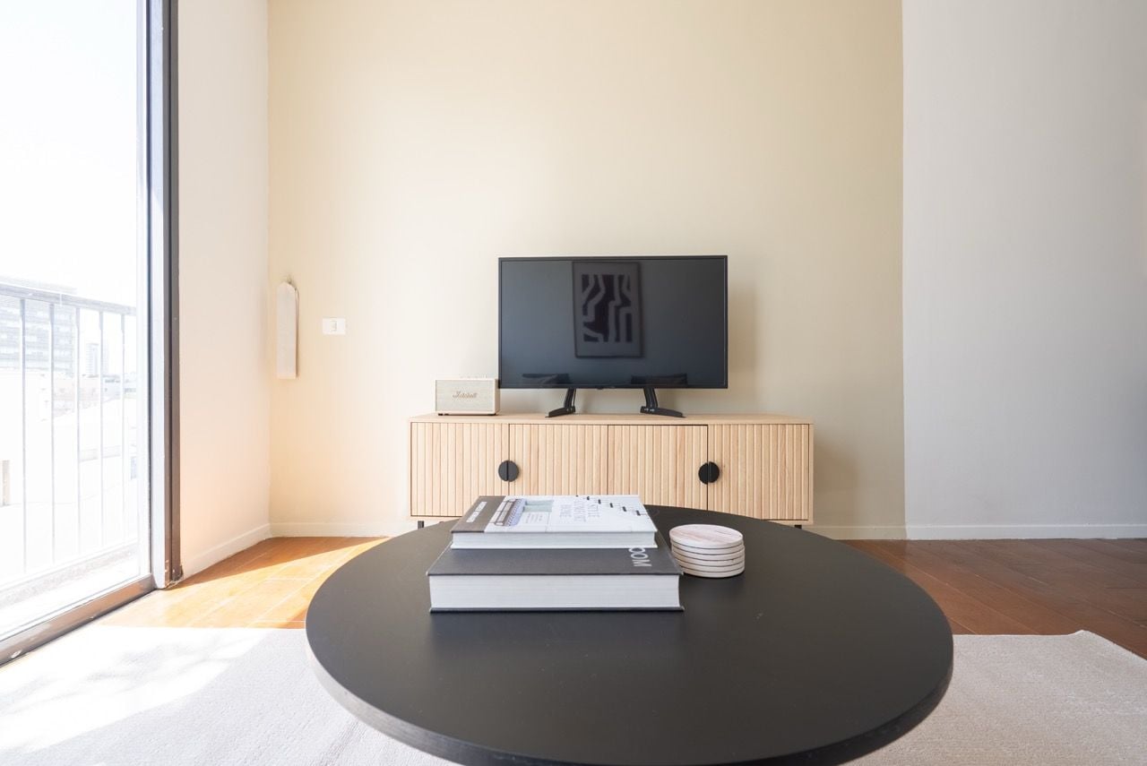 Ein modernes Wohnzimmer mit einem runden schwarzen Tisch, einem Flachbildfernseher auf einer Holzkommode und Blick durch ein großes Fenster.