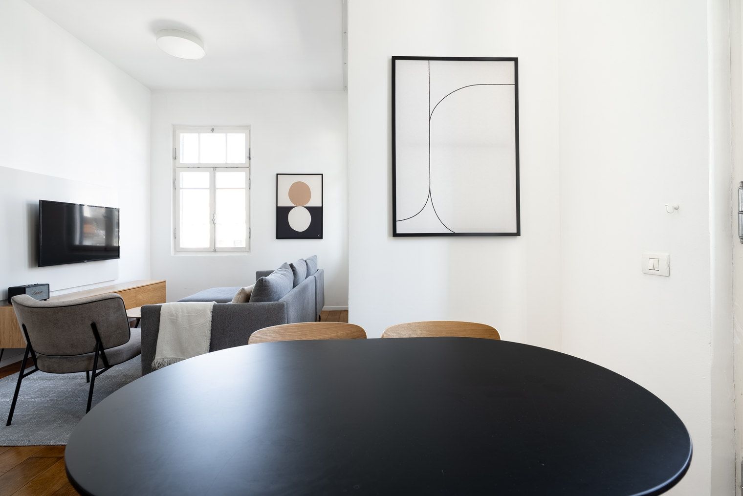 Una sala de estar moderna con una mesa redonda negra, un sofá gris y un televisor montado en la pared.