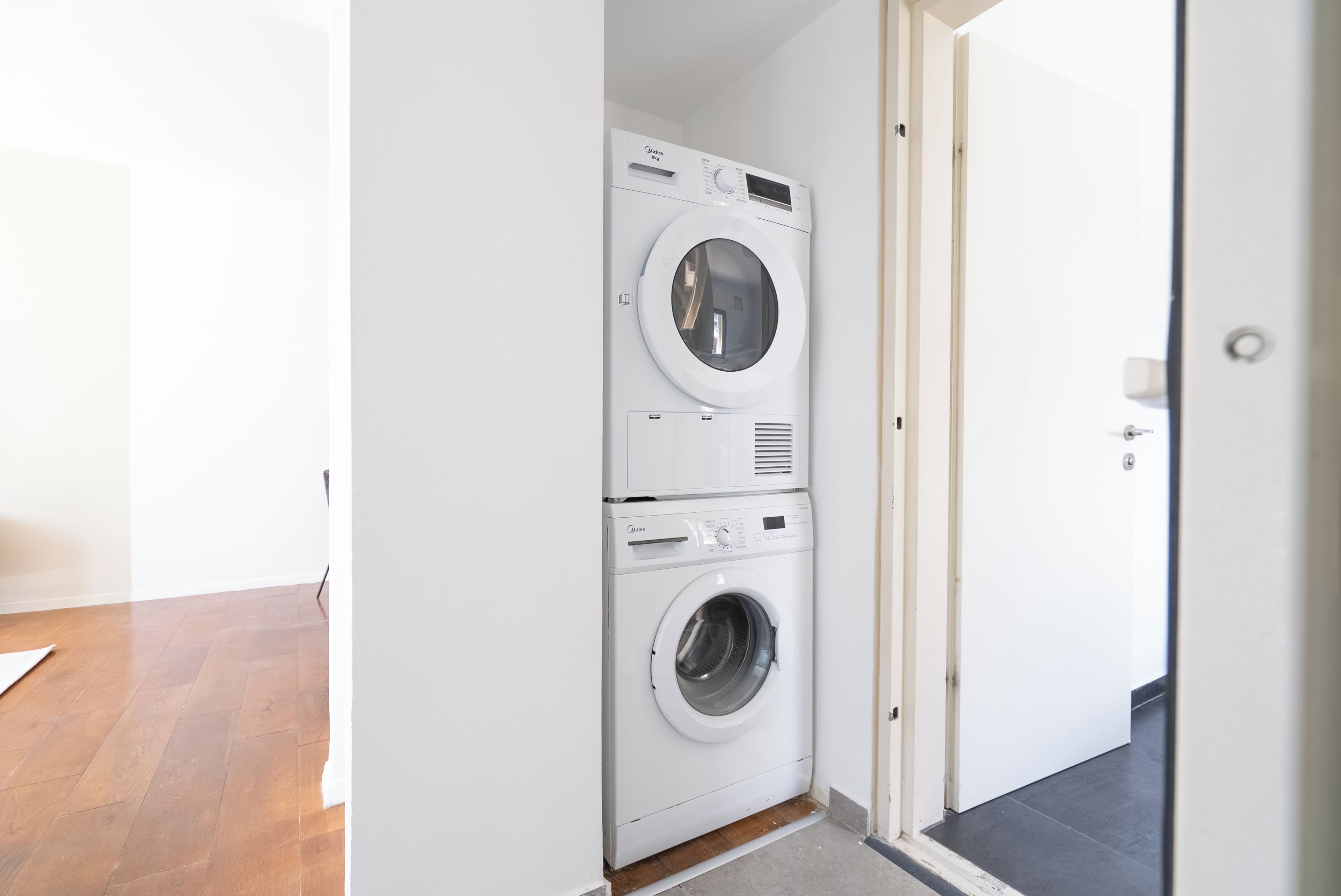 Een kleine wasruimte met een gestapelde wasmachine en droger, een houten vloer en een deur naar een andere kamer.