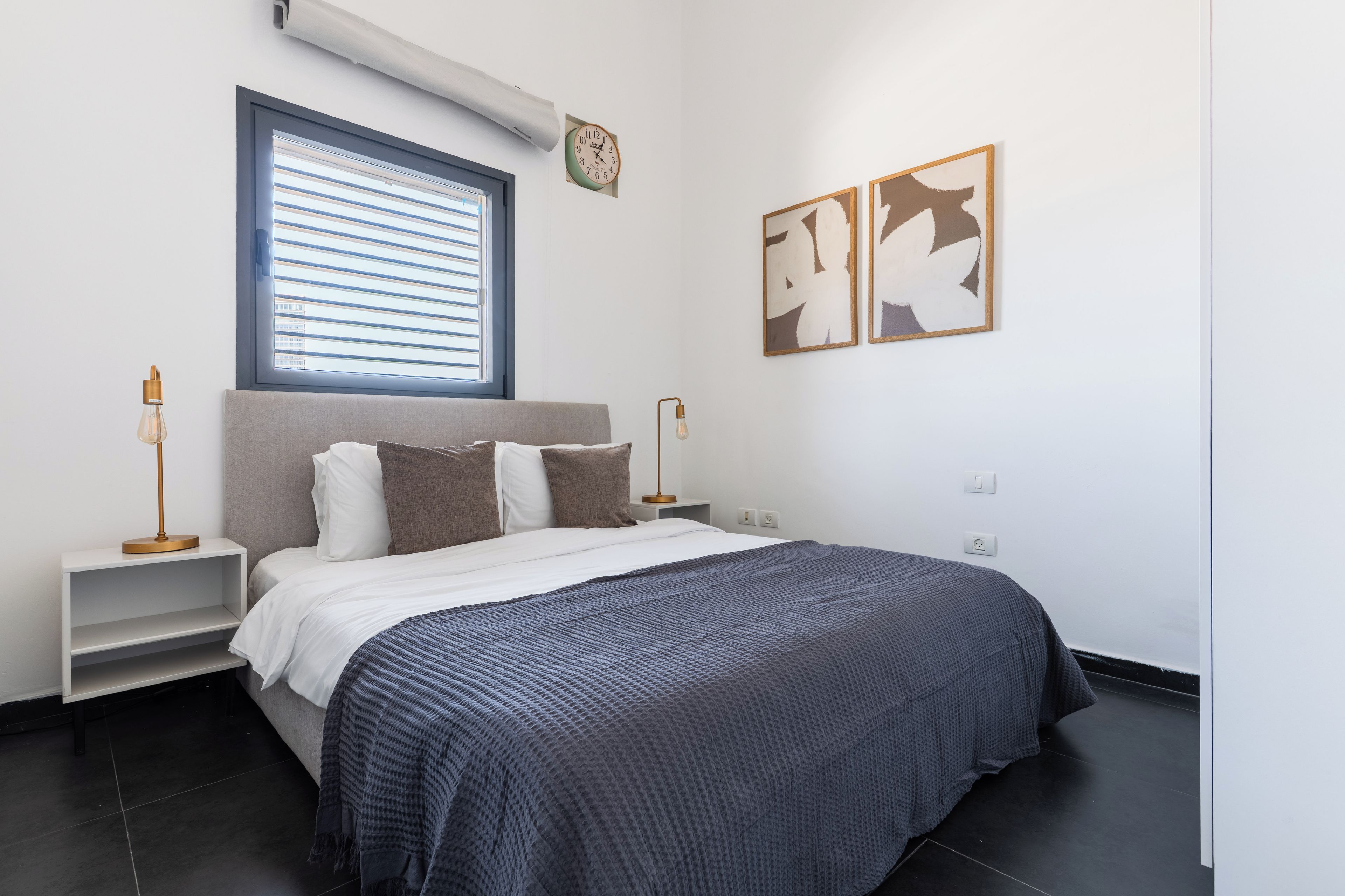Een moderne slaapkamer met een bed, nachtkastjes, een raam met zonwering, een wandklok en abstracte kunst.