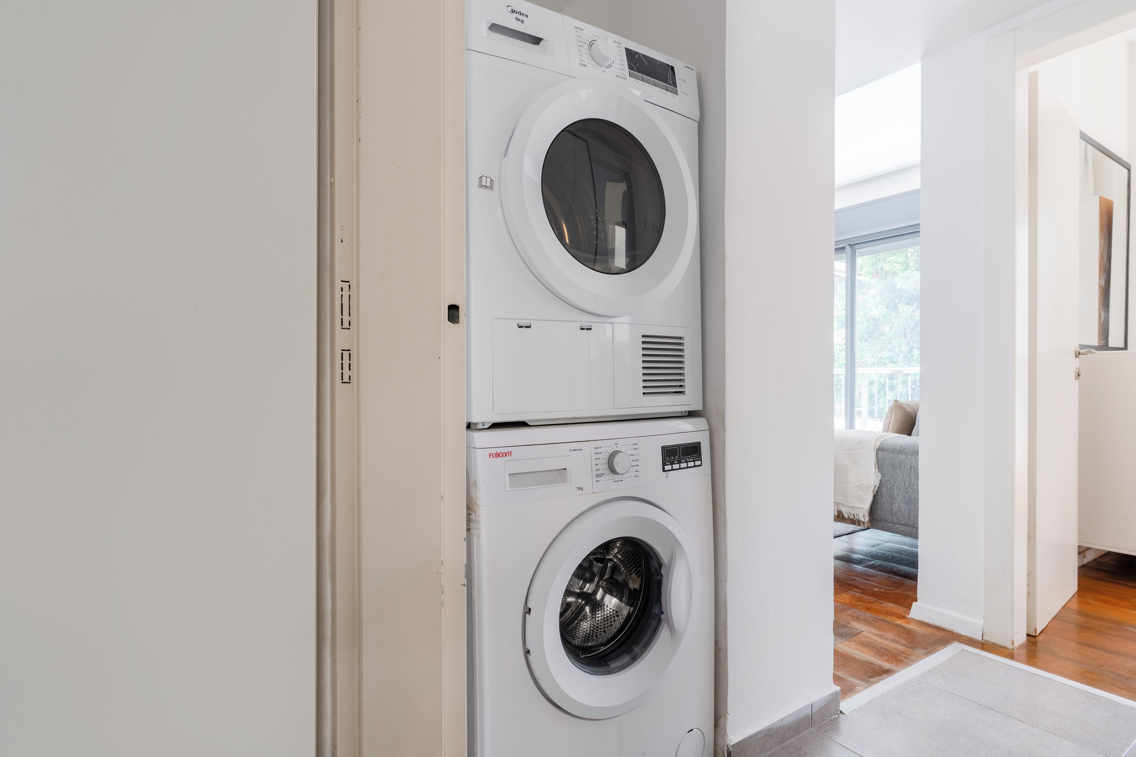 Un lavadero con dos lavadoras superpuestas, un panel de control y una puerta que da a una sala de estar con un sofá.