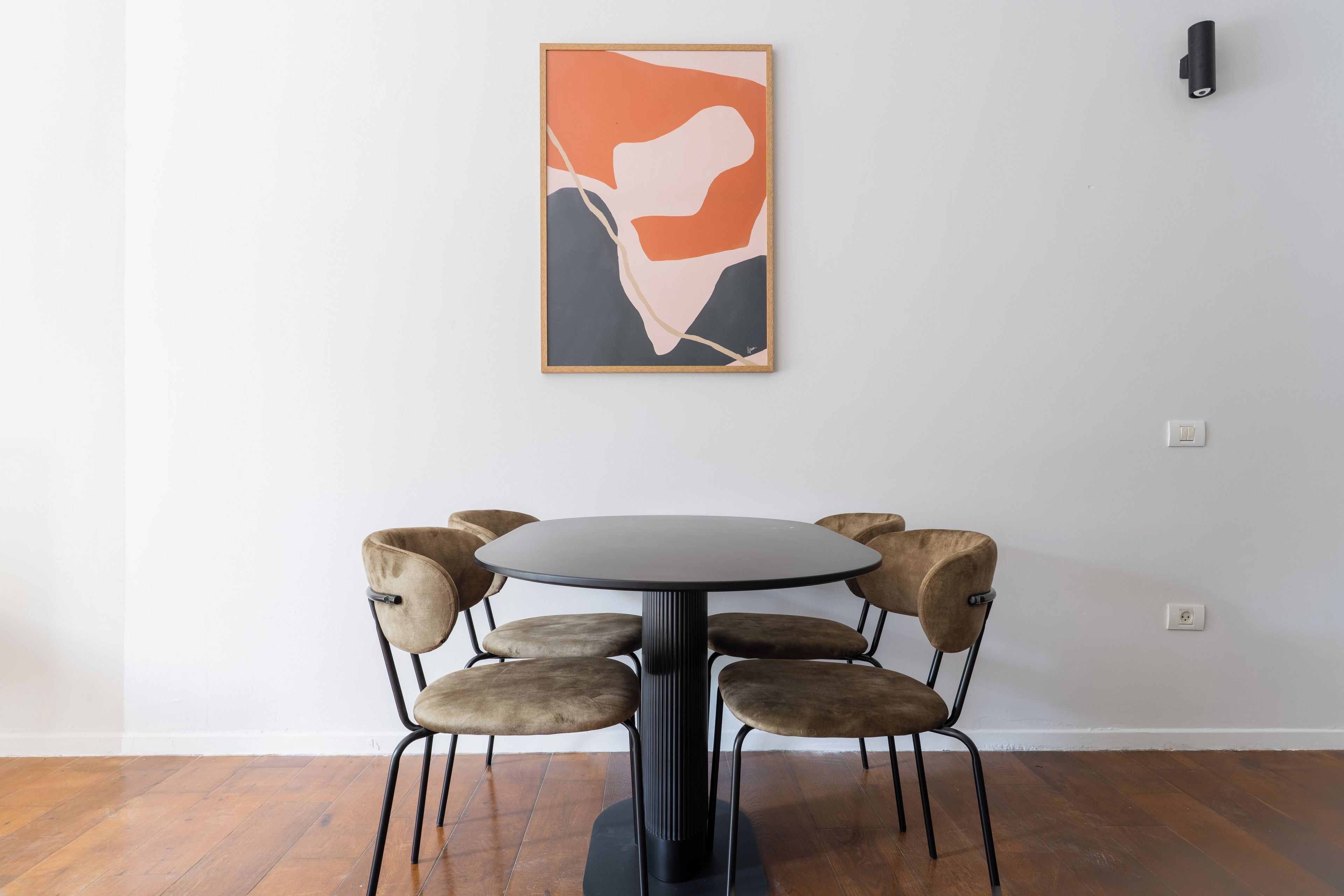 Un área de comedor con una mesa redonda y seis sillas, un cuadro abstracto enmarcado en la pared, y una lámpara de pared.