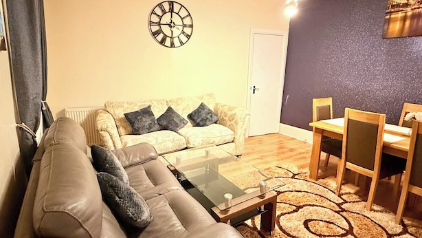 Ein Wohnzimmer mit einer Couch, einem Couchtisch, einem Esstisch mit Stühlen, einer Wanduhr und einem gemusterten Teppich.