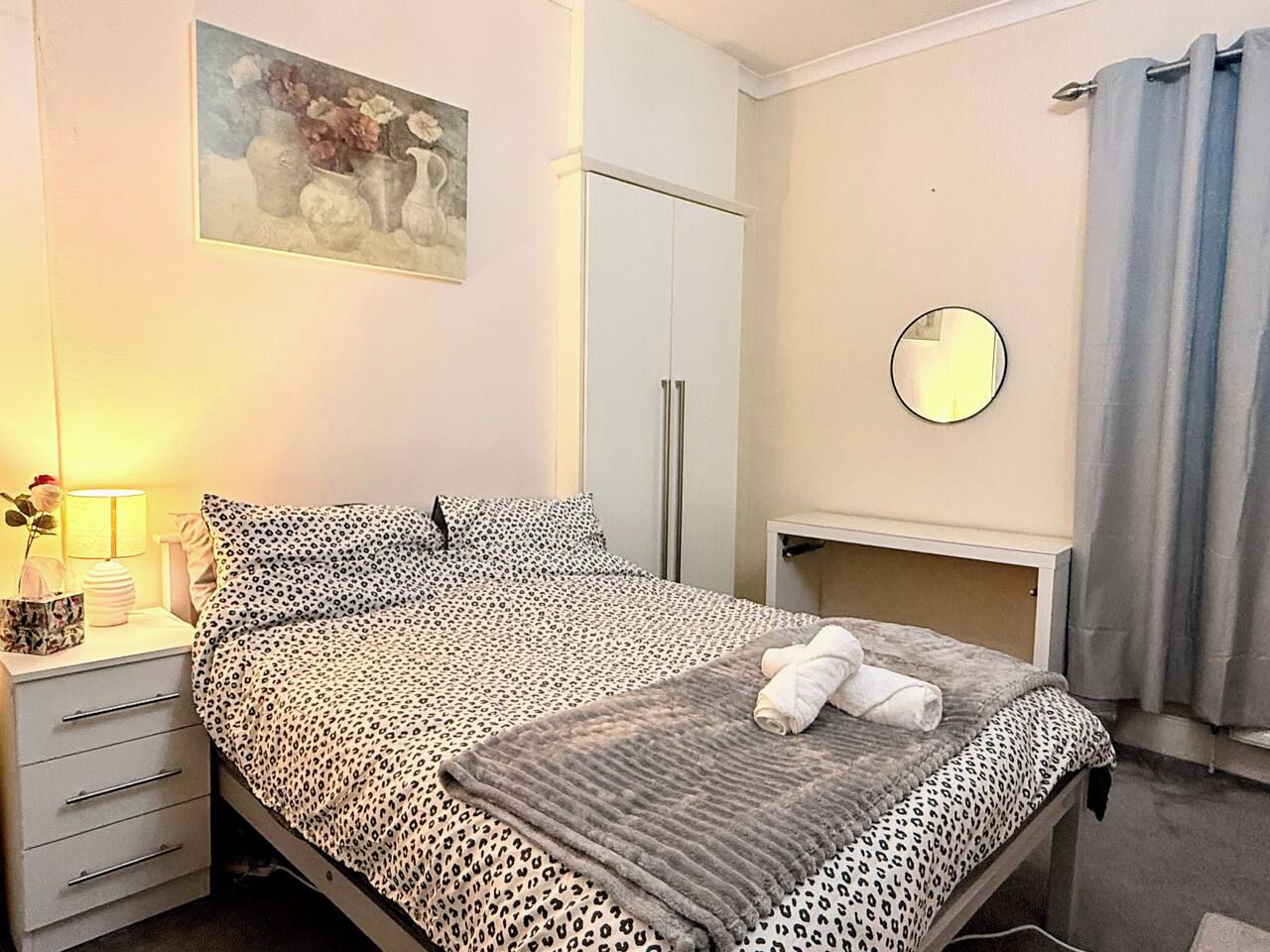 A neatly made bed with a leopard print bedspread, a bedside lamp, and a small table with a mirror.