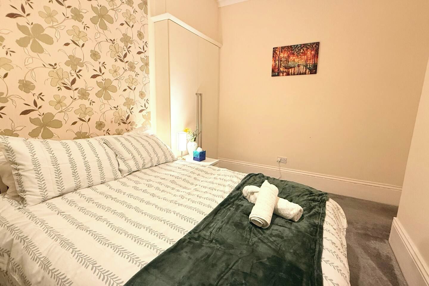 Un lit bien fait, avec une couvre-lit à rayures et une couverture vert foncé, un tableau accroché au mur, et une table de chevet sur laquelle se trouvent une lampe et un vase.