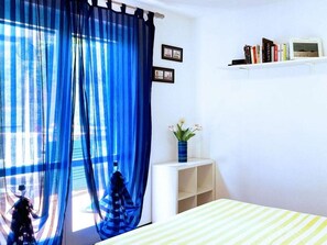Ein Zimmer mit einem Bett, einem Fenster mit blauen Vorhängen, einem kleinen weißen Nachttisch mit Blumen und einem Bücherregal mit Büchern.