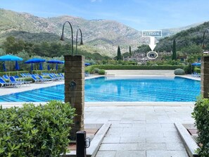 Ein Swimmingpool mit blauen Fliesen, umgeben von Liegestühlen und Sonnenschirmen, mit einer Bergkulisse im Hintergrund.
