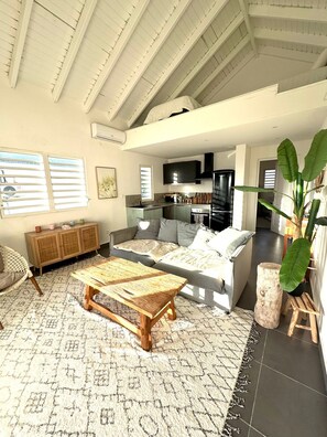 A spacious living room with a high ceiling, a sofa set, a coffee table, and a kitchen area visible in the background. - Petite Maison Duplex Cosy • Piscine • Près de Pinel (Cul-de-Sac)