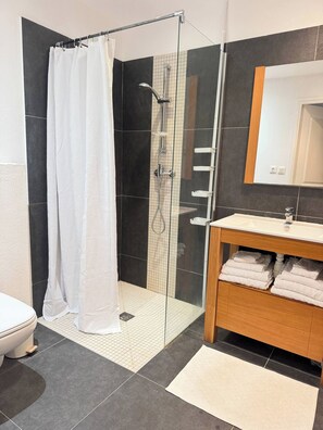 A modern bathroom with a glass-enclosed shower, a wooden vanity with a mirror, and a toilet. - Petite Maison Duplex Cosy • Piscine • Près de Pinel (Cul-de-Sac)