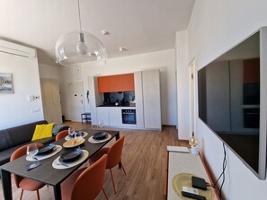 A modern kitchen with orange cabinets, a dining area with a table set for two, and a large TV on the wall.