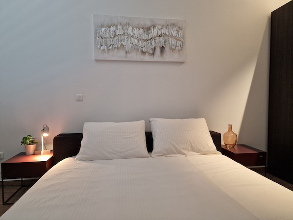 Un dormitorio con cama, dos mesitas de noche, una obra de arte en la pared, una lámpara de noche y una planta en maceta.