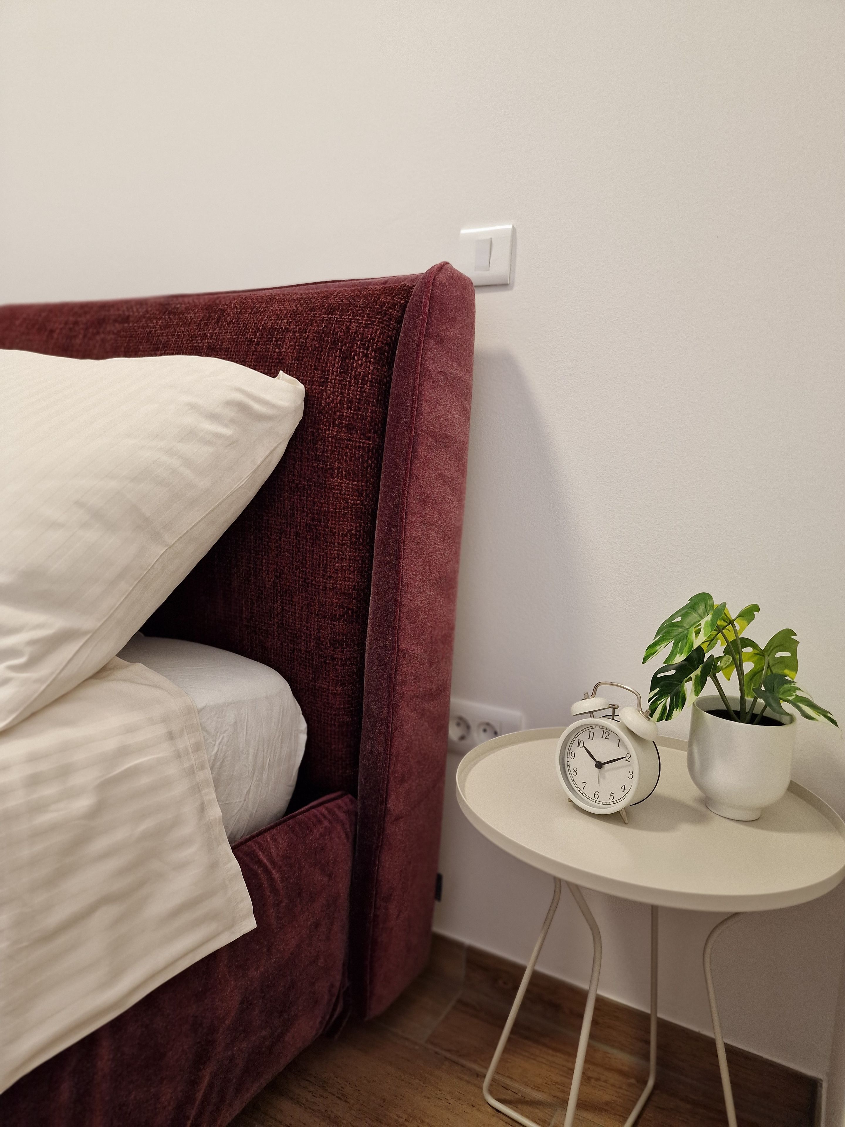 A bed with white bedding, a red headboard, a white alarm clock, and a potted plant on a small table.