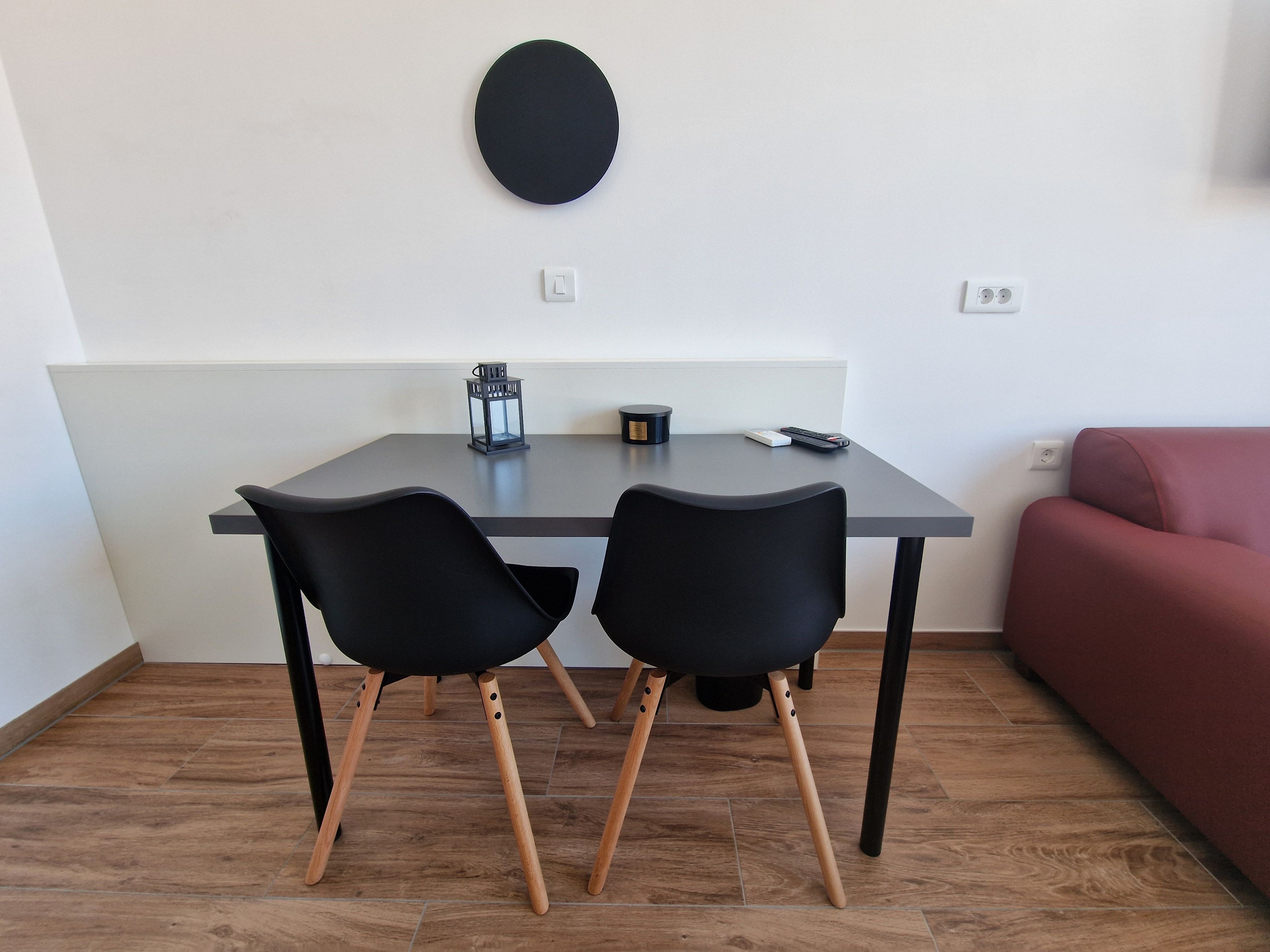 Ein kleiner, moderner Raum mit einem dunkelgrauen Tisch, zwei schwarzen Stühlen, einem roten Sofa und einer runden Wanddekoration.