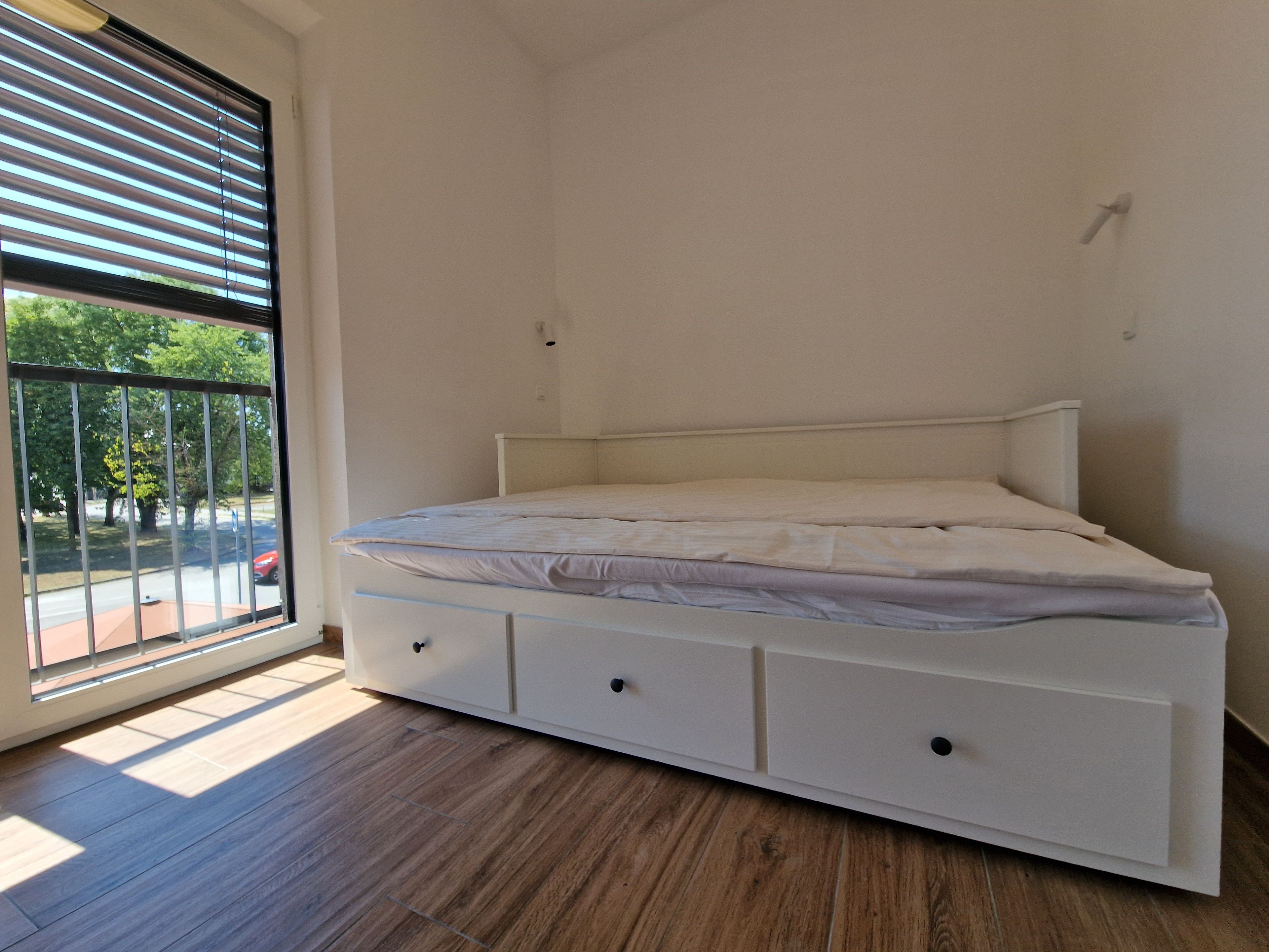 Habitación individual con una cama y cabecera blanca, ventana con persianas y vistas al exterior.