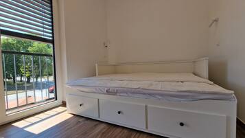 Habitación individual con una cama y cabecera blanca, ventana con persianas y vistas al exterior.