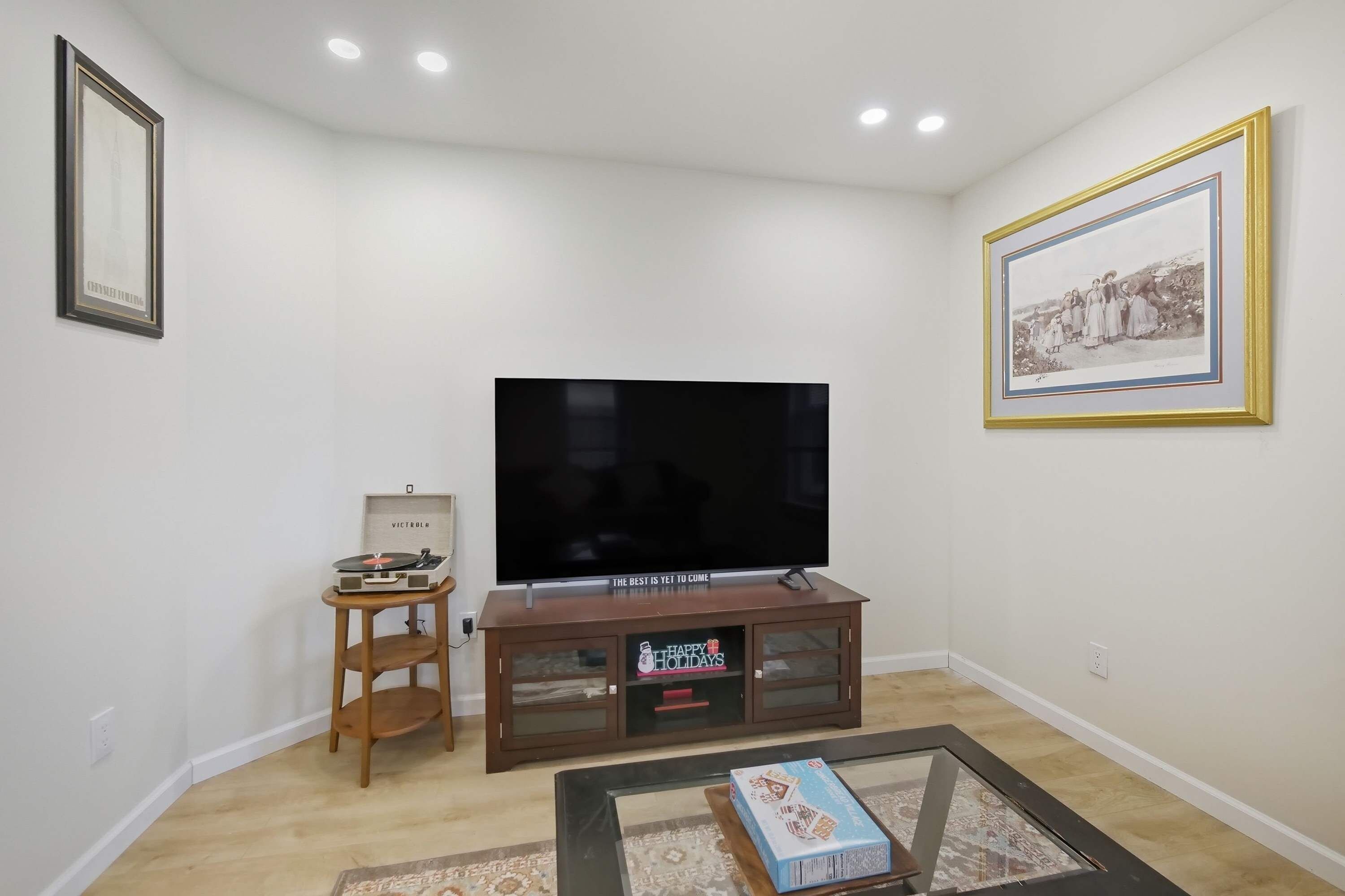 Una sala de estar con un televisor de pantalla plana sobre un soporte de madera, una mesita con un libro y un cuadro en la pared.