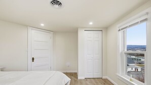 Quarto com cama, duas portas brancas, janela com vista e um respiradouro no teto.