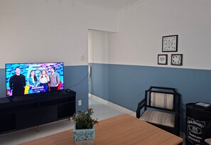A living room with a television displaying a show, a wooden dining table, a black sofa, and a Jack Daniel's whiskey barrel. - SHB - Aconchego a Passos do Mar (Santos)