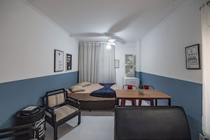 Una habitación pequeña y funcional con cama, escritorio, silla y una mesita. Cuenta con ventilador de techo, ventana con cortinas y repisas con adornos.
