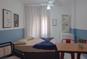Un dormitorio con cama, escritorio, silla, ventilador de techo y ventana con cortinas.