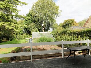 Eine Holzterrasse mit einem Metalltisch, umgeben von einem weißen Zaun, mit Blick auf einen Garten mit Gewächshaus und verschiedenen Bäumen.