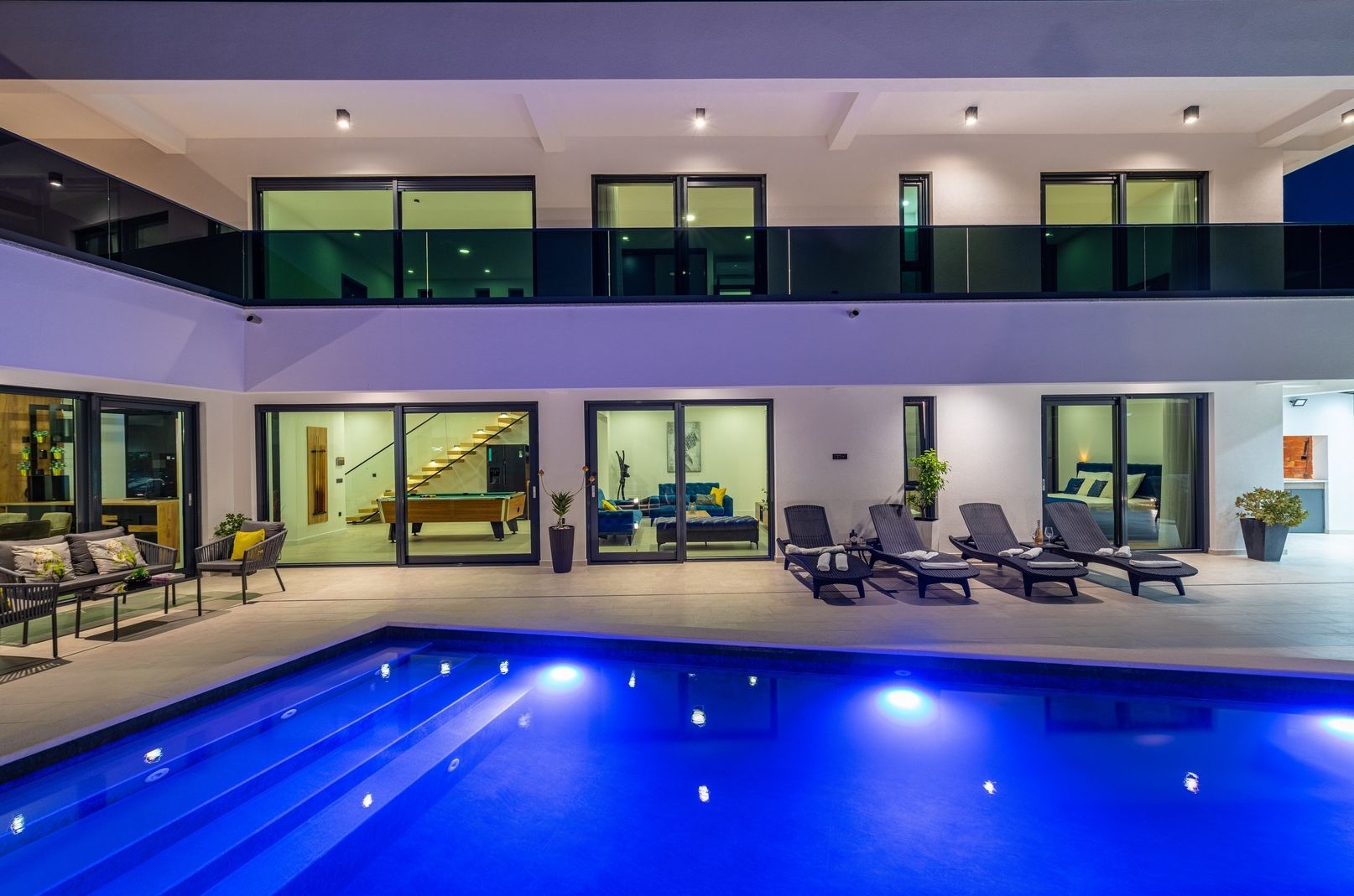 A modern indoor pool area with lounge chairs, a staircase, and a living room with a sofa and coffee table.