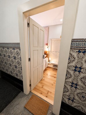 Een gang met een houten vloer, een deur die leidt naar een slaapkamer, en behang met een patroon.
