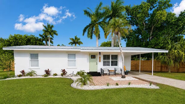 A single-story house with a covered porch, a small front yard with a lawn and potted plants, and a wooden fence in the background. - The McKenzie House (Punta Gorda)