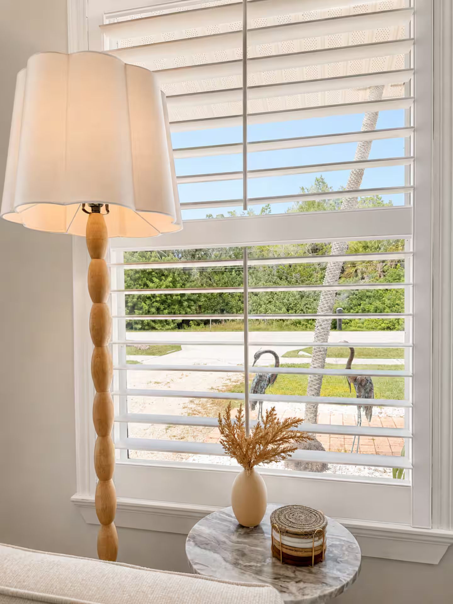 Una habitación con ventana y persianas horizontales, una lámpara de pie de madera, una mesa de mármol con un jarrón y un objeto decorativo, y vistas a un jardín con árboles y un camino.