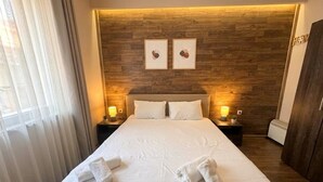 Une chambre d’hôtel avec une tête de lit en bois, un lit recouvert de draps blancs, deux lampes de chevet et une fenêtre ornée de rideaux légers.