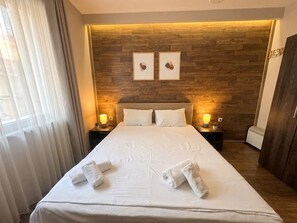 Una habitación de hotel con cabecera de madera, cama con ropa blanca, dos lámparas de noche y una ventana con cortinas traslúcidas.