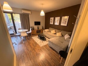 Una sala moderna con sofá, mesa de centro y televisor. Hay un balcón con mesa y sillas afuera.