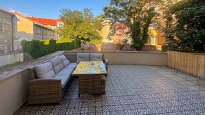 Une terrasse extérieure avec un ensemble de canapés et une table, entourée d’un sol à motifs et offrant une vue sur des bâtiments et des arbres.
