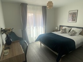 A hotel room with a bed, a desk, a chair, a vase with flowers, a lamp, and a framed picture on the wall.