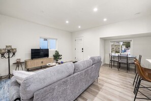 Ein modernes Wohnzimmer mit einem grauen Sofa, einem Fernseher auf einer Holzkommode, einem Esstisch mit Stühlen und einem Fenster mit Jalousien.