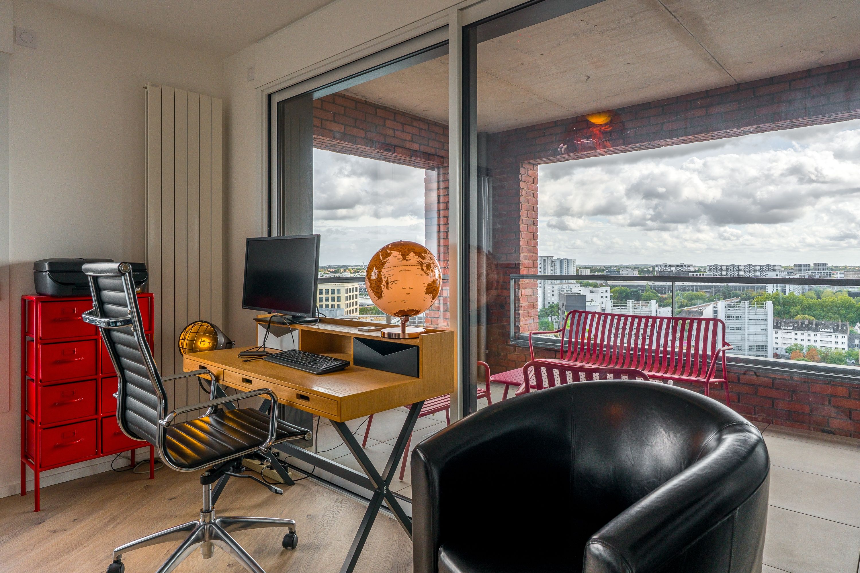 A modern office space with a desk, computer, and a globe. There is a red chair and a black leather chair. The room has large windows with a view of buildings.