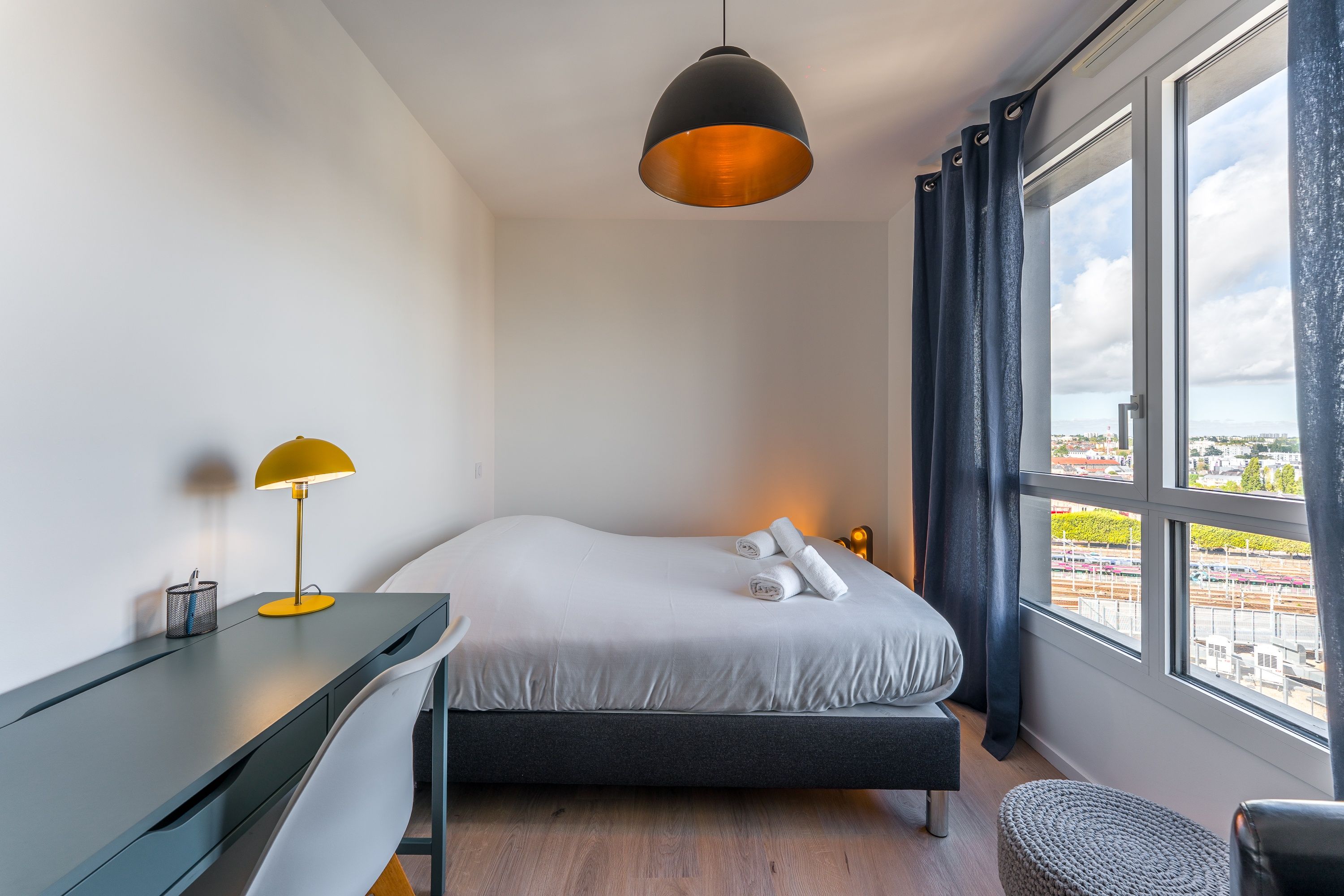Een hotelkamer met een bed, een bureau, een lamp en uitzicht op de stad.
