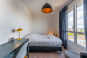 Een hotelkamer met een bed, een bureau, een lamp en uitzicht op de stad.