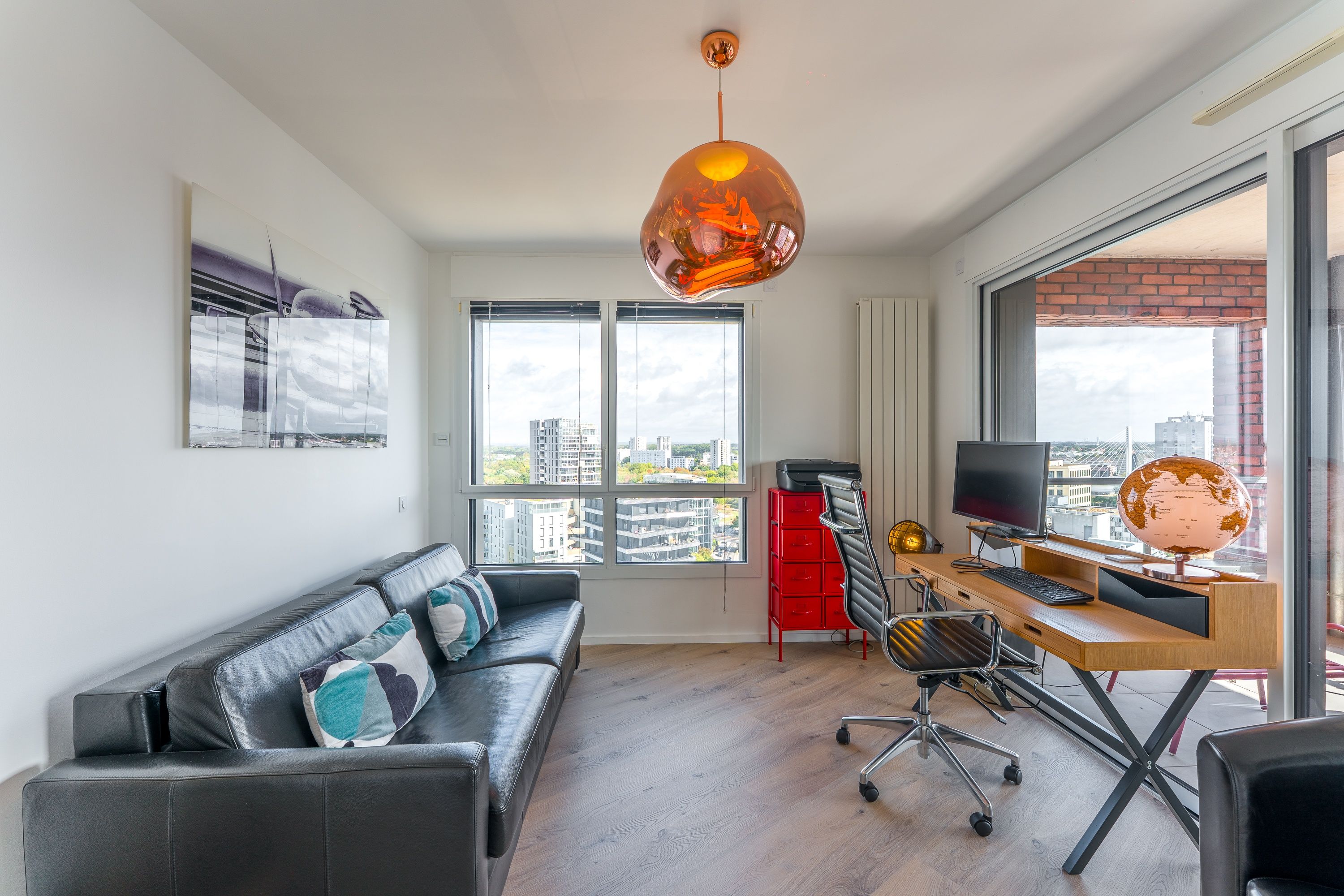 A modern living room with a leather sofa, a wooden desk with a computer, and a large window offering a city view.