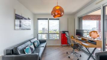 A modern living room with a leather sofa, a wooden desk with a computer, and a large window offering a city view.