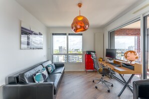 Een moderne woonkamer met een leren bank, een houten bureau met een computer en een groot raam met uitzicht op de stad.