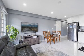 Una sala moderna con un sofá, una mesa de comedor con sillas, un televisor de pantalla plana montado en la pared y una cocina con refrigerador.