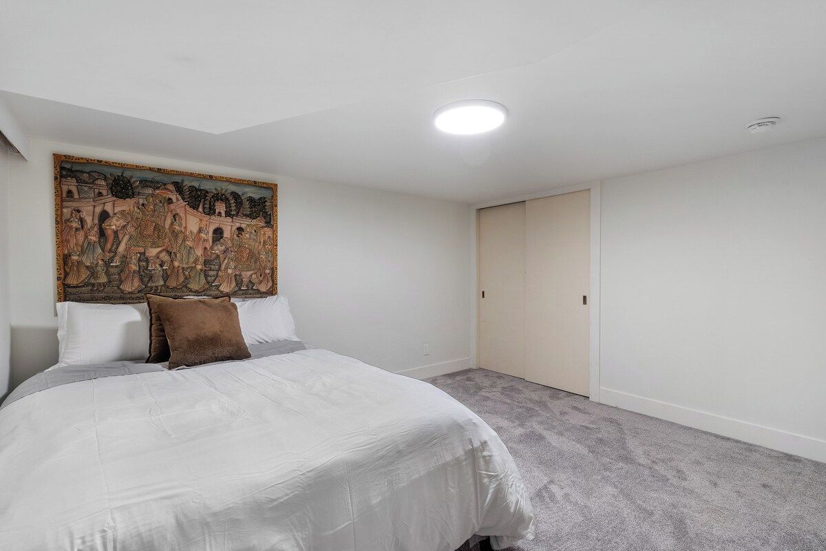 A bedroom with a bed, a headboard with a painting, a wardrobe, and a carpeted floor.