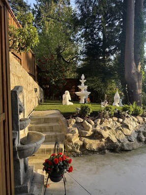 Eine Steintreppe führt zu einer Holztür, einem Steinfountain und einem Garten mit Statuen und roten Blumen.