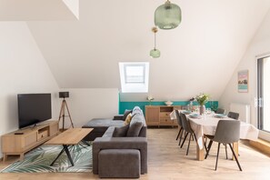 A modern loft apartment with a dining area, a living room with a sofa and TV, and a kitchen with a window. - Loft au Vert – 92 m², balcon & proche plages (Saint-Lunaire)