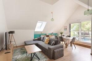A modern living room with a sectional sofa, dining table, and chairs. There is a large window providing natural light and a view of greenery outside. - Loft au Vert – 92 m², balcon & proche plages (Saint-Lunaire)